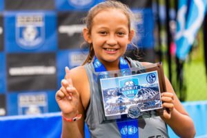 Young Kick It 3v3 Soccer player holding a National Championship plaque and medal while posing with a number one gesture at a tournament event