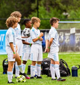 Youth soccer players in Kick It 3v3 Soccer uniforms standing on the field during a tournament, preparing for play with soccer balls and gear nearby