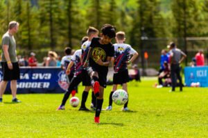 Youth soccer player kicking a soccer ball during a Kick It 3v3 Soccer tournament while other players and spectators stand in the background on a sunny outdoor field