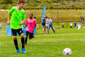 Youth players compete during a Kick It 3v3 Soccer tournament match, chasing the ball across a grass field during fast-paced small-sided play