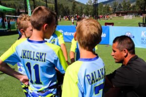 Youth soccer players in Kick It 3v3 jerseys gather around a coach during a team huddle on an outdoor soccer field, with other games and mountain scenery visible in the background