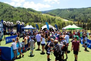 Players, families, and spectators gathered at a Kick It 3v3 Soccer tournament in a mountain setting, with soccer activities, tents, and scenic hills in the background