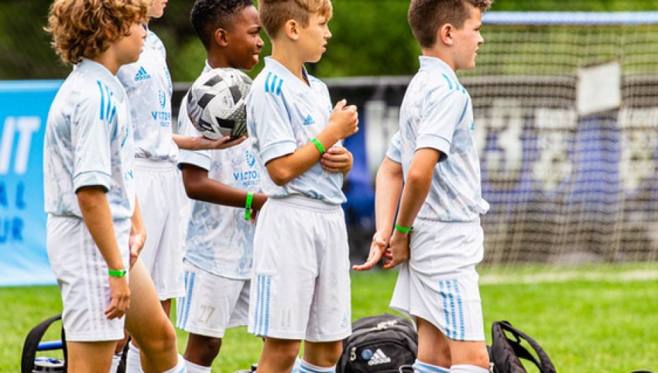 Youth soccer players in Kick It 3v3 Soccer uniforms standing on the field during a tournament, preparing for play with soccer balls and gear nearby
