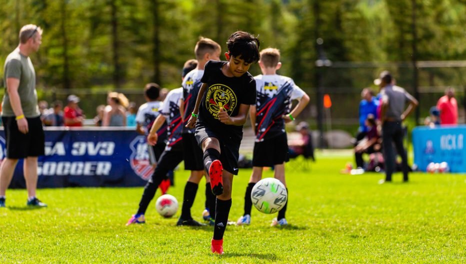 Youth soccer player kicking a soccer ball during a Kick It 3v3 Soccer tournament while other players and spectators stand in the background on a sunny outdoor field