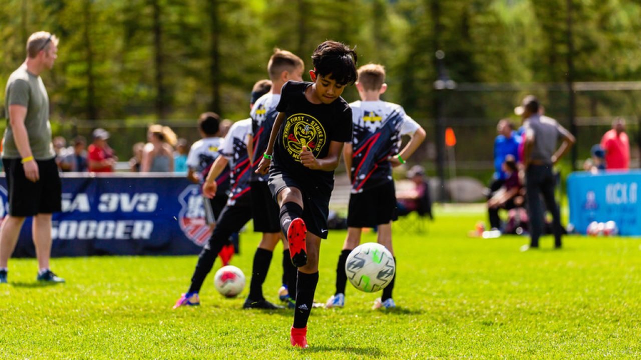 Youth soccer player kicking a soccer ball during a Kick It 3v3 Soccer tournament while other players and spectators stand in the background on a sunny outdoor field