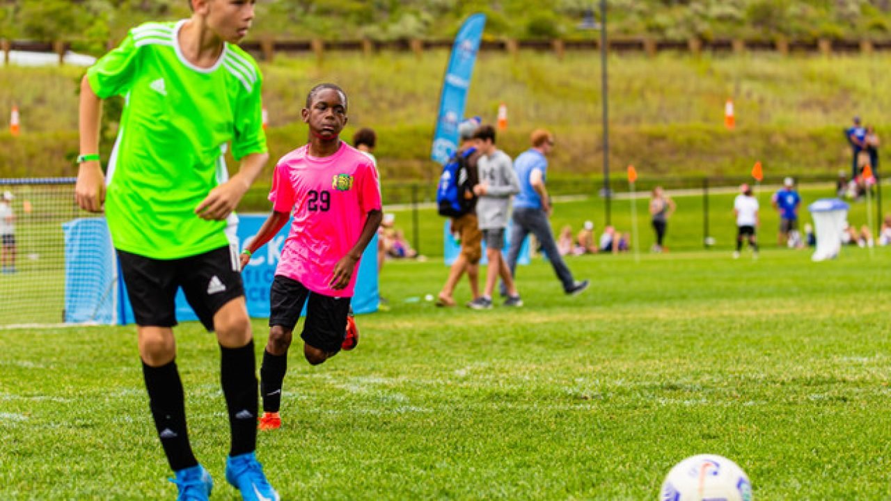 Youth players compete during a Kick It 3v3 Soccer tournament match, chasing the ball across a grass field during fast-paced small-sided play