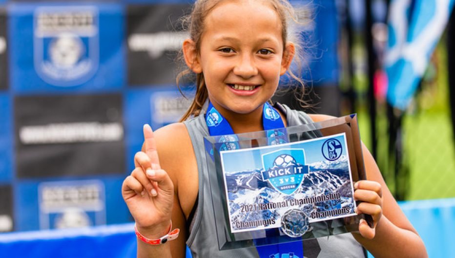Young Kick It 3v3 Soccer player holding a National Championship plaque and medal while posing with a number one gesture at a tournament event