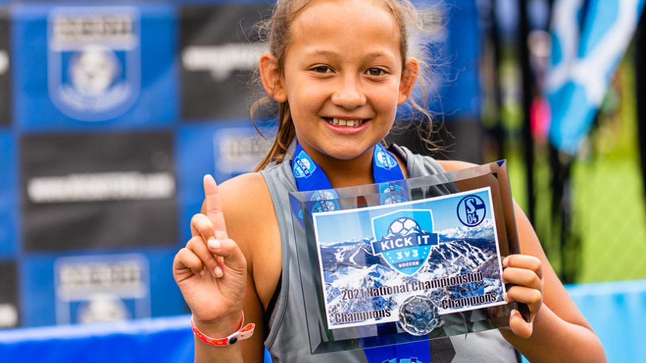 Young Kick It 3v3 Soccer player holding a National Championship plaque and medal while posing with a number one gesture at a tournament event