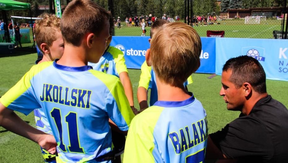 Youth soccer players in Kick It 3v3 jerseys gather around a coach during a team huddle on an outdoor soccer field, with other games and mountain scenery visible in the background