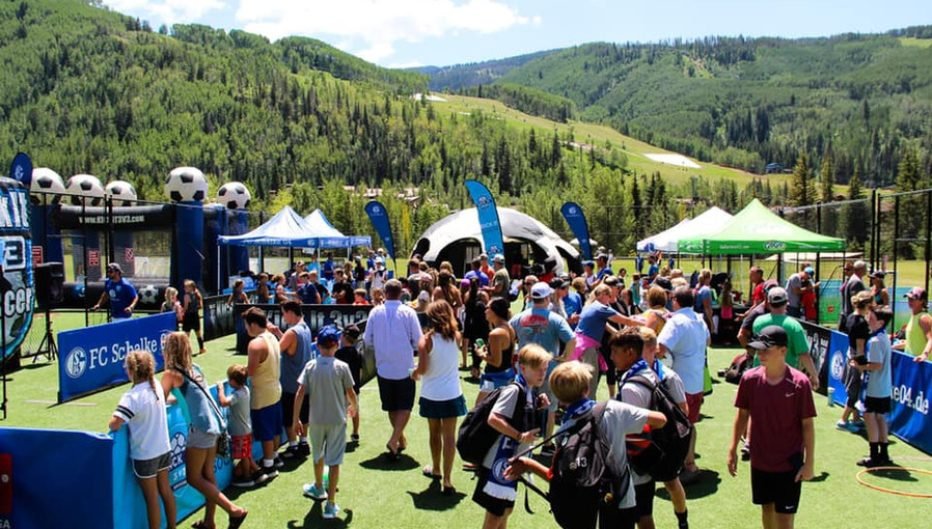Players, families, and spectators gathered at a Kick It 3v3 Soccer tournament in a mountain setting, with soccer activities, tents, and scenic hills in the background