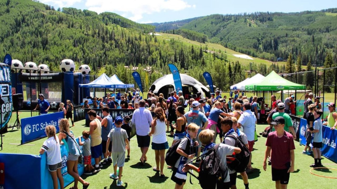 Players, families, and spectators gathered at a Kick It 3v3 Soccer tournament in a mountain setting, with soccer activities, tents, and scenic hills in the background