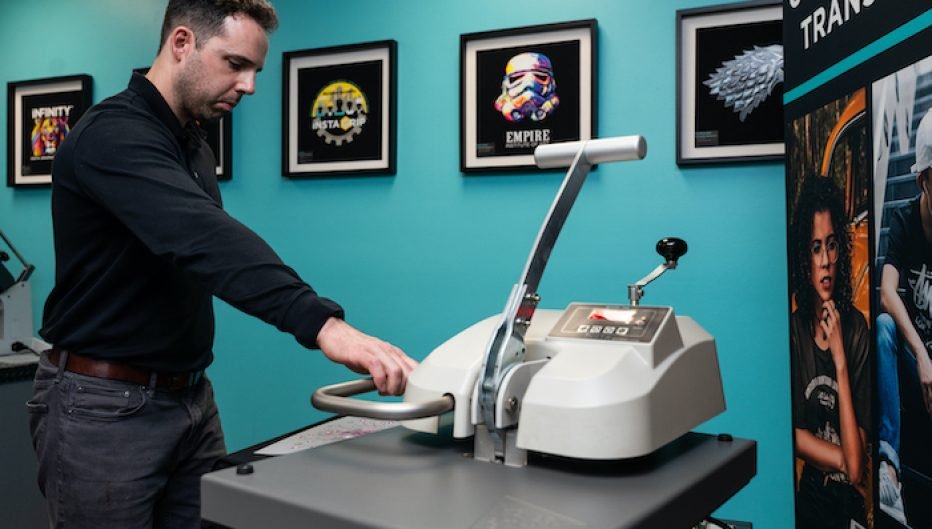 Technician operating an Insta Graphic Systems heat press machine in a showroom, demonstrating custom heat transfer application technology with branded transfer samples displayed on the wall